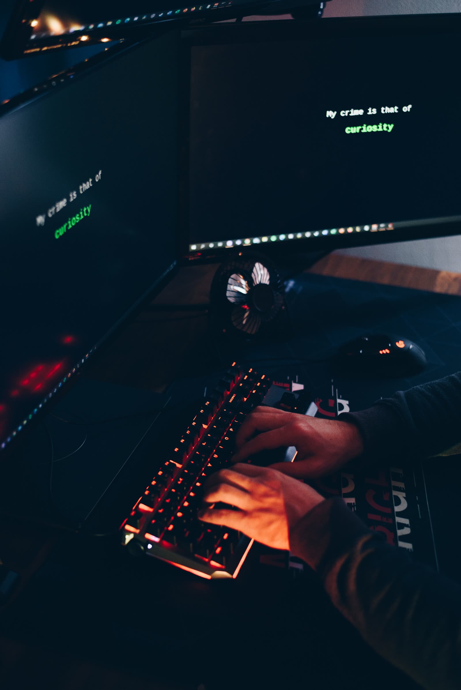 A dark corner desk with dual monitors and a red-backlit mechanical keyboard glowing under a player's hands