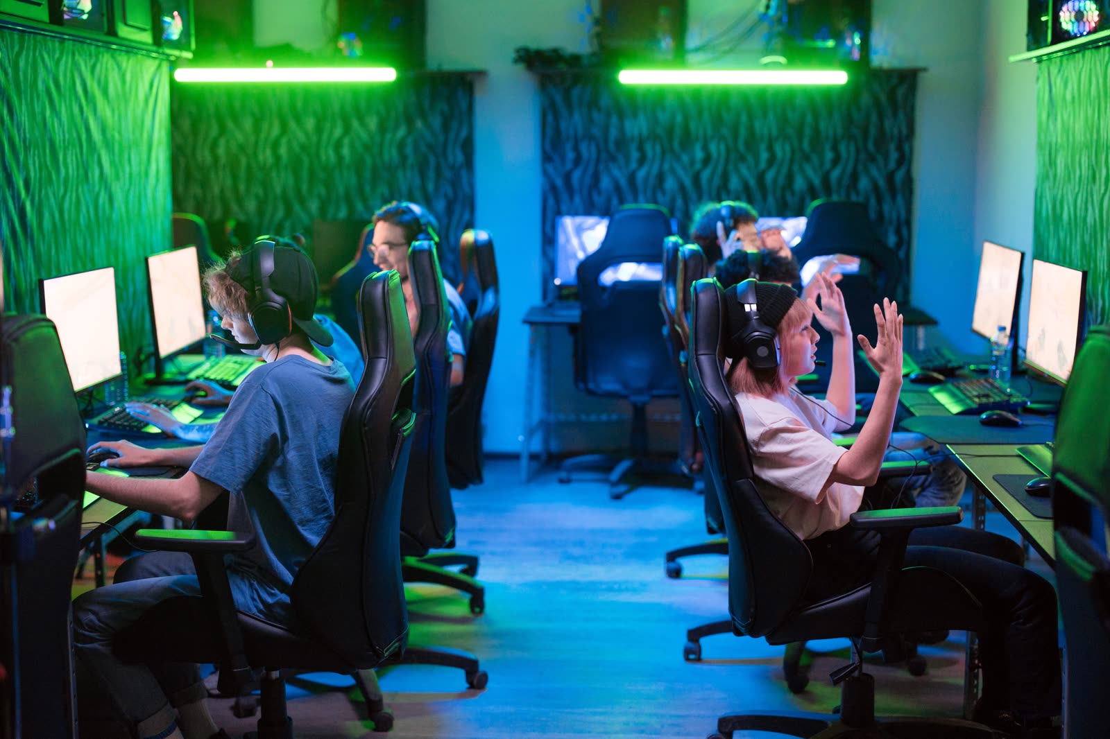 A wide gaming room bathed in green light with two rows of gaming chairs facing screens along both walls