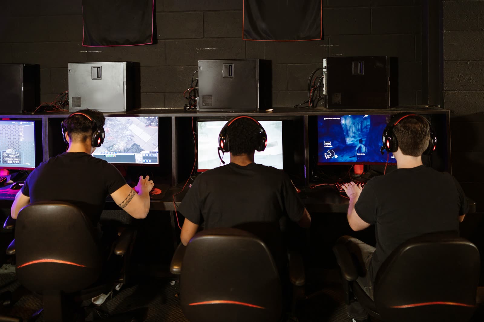 Three players seen from behind, side by side at their monitors in a dark room, headsets on