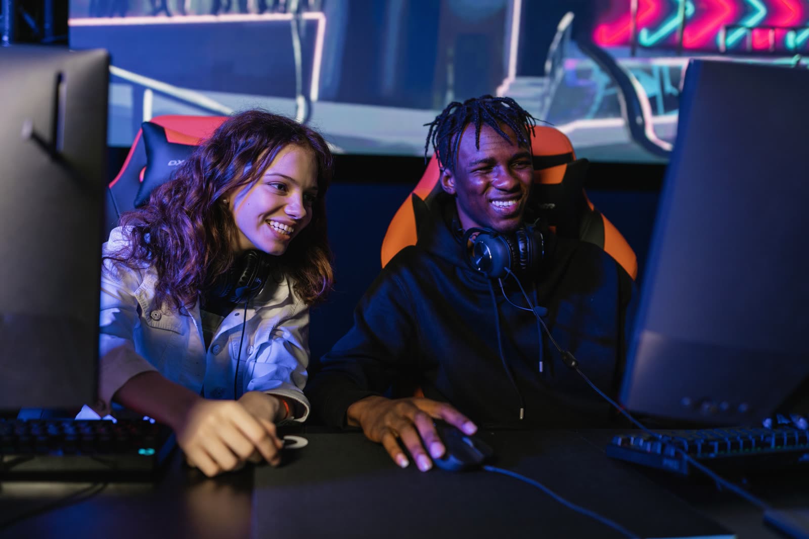 Two smiling players in orange gaming chairs at neighbouring stations, neon lights glowing behind them